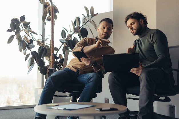 Colegas do sexo masculino discutindo brainstorming de projetos de negócios para ideias usando computadores portáteis no escritório moderno