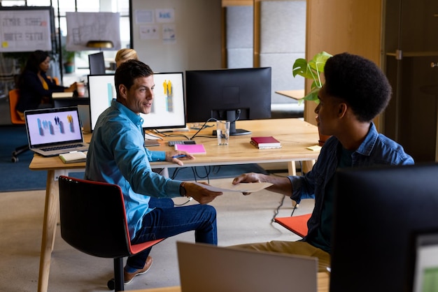 Colegas masculinos felizes e diversificados nas mesas, passando documentos no escritório, escritório casual, trabalho em equipa, negócios e trabalho criativo, inalterados.