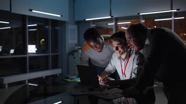 Foto colegas multiétnicos trabajan en una laptop y sonríen en una oficina oscura