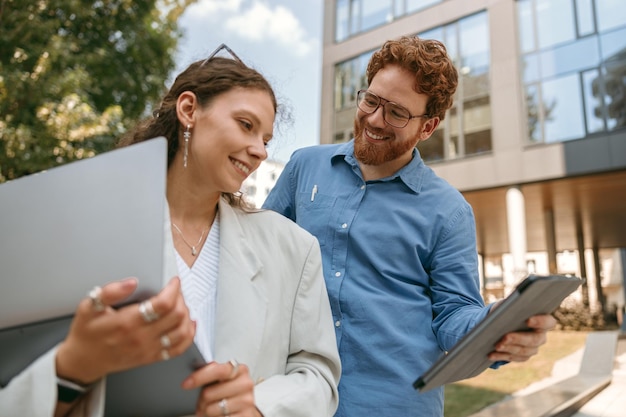 Colegas de negocios sonrientes discuten problemas comerciales mientras usan una computadora portátil de pie en el fondo del edificio