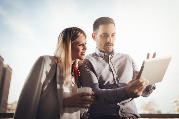 Colegas de negocios tomando café y usando una tableta digital en el balcón frente a la oficina.