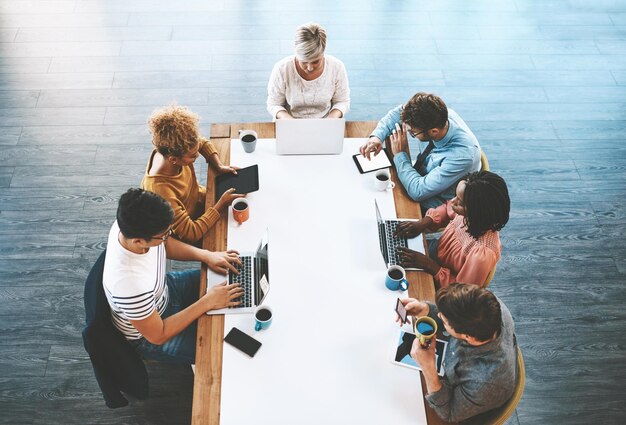 Colegas que planean discutir estrategias y buscar en línea Diversos empresarios hablando en una reunión escribiendo en computadoras portátiles y buscando en tabletas ideas juntos en una oficina en el trabajo desde arriba