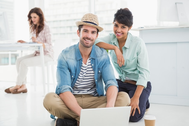 Colegas sonrientes que se sientan en el piso usando la computadora portátil