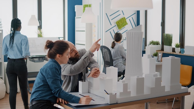 Colegas usando smartphone para analisar o modelo de construção e o plano de projetos na mesa. Homem e mulher trabalhando com telefone celular para projetar a estrutura da construção. Projeto de desenvolvimento urbano