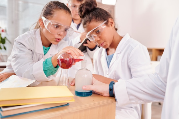 Colegiais maravilhadas com óculos de proteção e jalecos brancos olhando para uma substância líquida azul em um bico grande, segurado pelo professor de química na aula