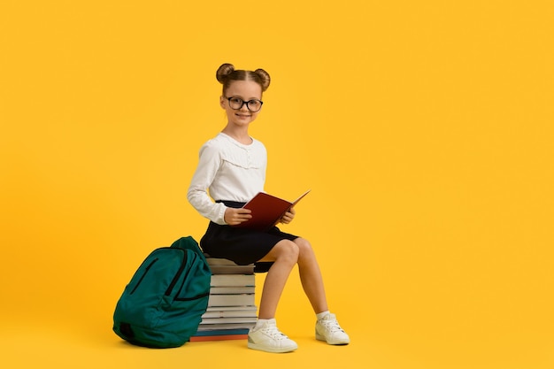 Colegial bonita em óculos sentado na pilha de livros sobre fundo amarelo