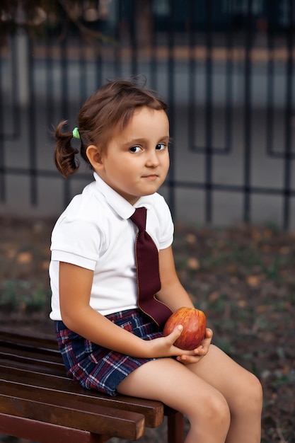 Colegial bonitinha de uniforme sentado em um banco e comendo uma maçã.