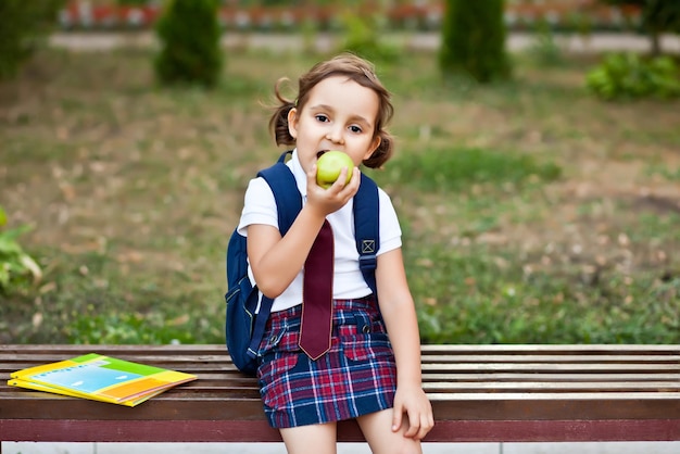 Colegial bonitinha de uniforme sentado em um banco e comendo uma maçã.