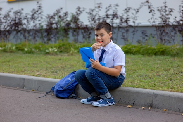 El colegial con una camisa blanca con una corbata azul, sostiene una lonchera azul y come un trozo de manzana