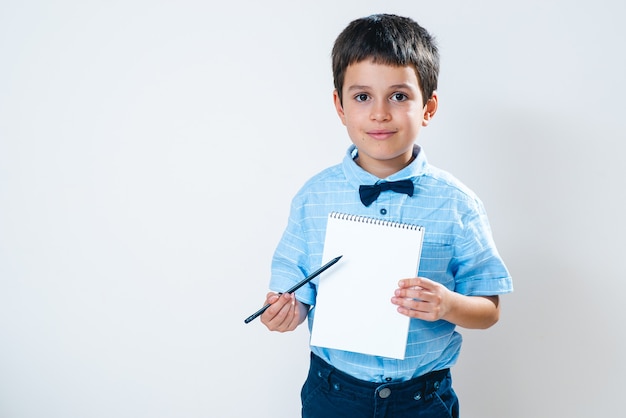 El colegial con una camisa con una pajarita sostiene un cuaderno frente a él y lo señala con un lápiz