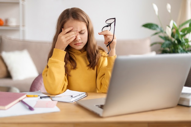 Colegial cansada esfregando os olhos, segurando os óculos, exausta de estudar em casa e fazer o dever de casa