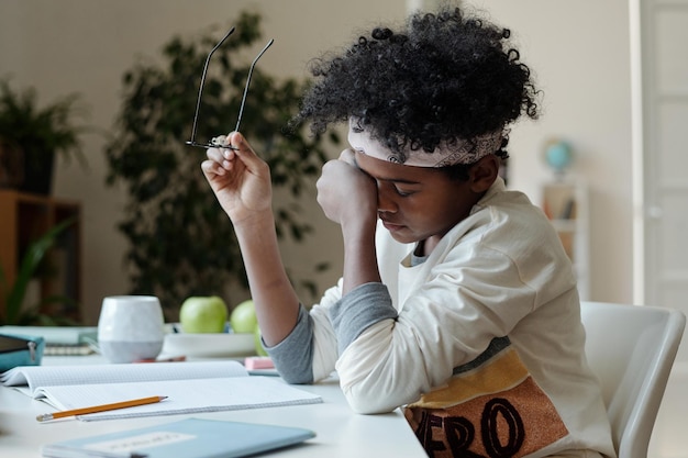 Colegial cansado frotándose los ojos mientras está sentado en el escritorio con cuaderno y lápiz