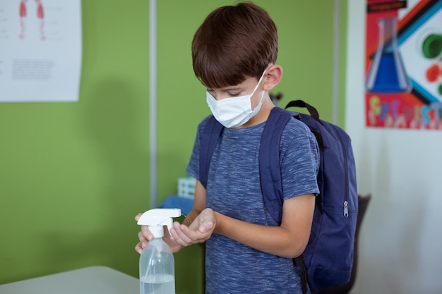 Colegial caucásico en el aula con mascarilla y mochilas escolares desinfectando las manos