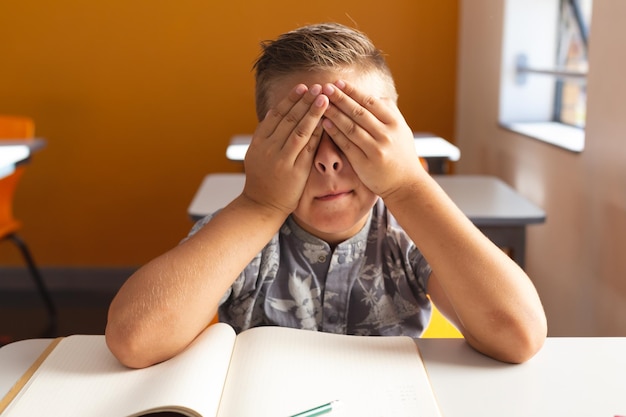 Colegial caucásico cansado sentado en el escritorio en el aula con un libro que cubre los ojos con las manos