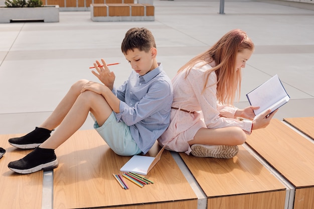 El colegial y la colegiala sentados espalda con espalda en un banco en el patio de la escuela cerca del edificio de la escuela. Estudiar con libros y cuadernos