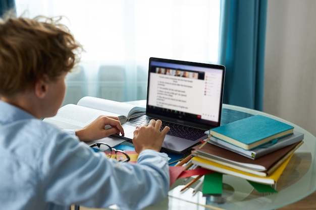 El colegial está escribiendo informe en la computadora portátil, haciendo los deberes