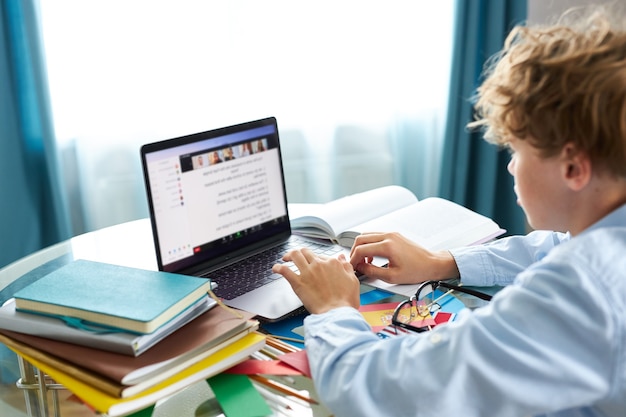 El colegial está escribiendo informe en la computadora portátil, haciendo los deberes