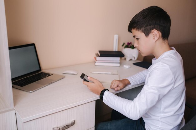 El colegial estudia en el escritorio de su habitación. El niño usa una computadora portátil y un teléfono inteligente para escribir en un cuaderno. Libros y t.