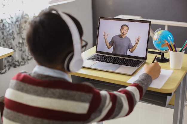 Foto un colegial está estudiando y mirando a su maestro en una computadora portátil mientras escribe en un papel