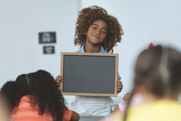 Colegial de etnia africana sosteniendo una pizarra y mirando a la cámara en el aula