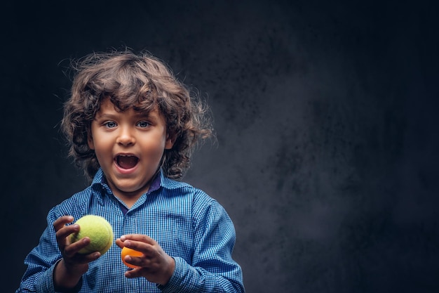 Un colegial feliz con cabello castaño rizado vestido con una camisa azul sostiene pelotas de tenis, diviértete en un estudio. Aislado en el fondo oscuro con textura.