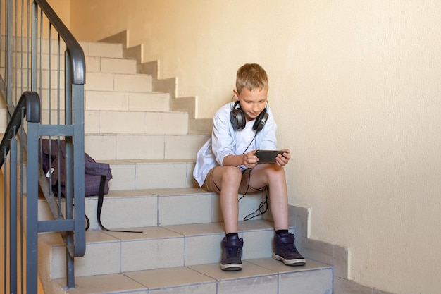 Un colegial feliz está sentado en las escaleras con auriculares y mirando el teléfono saltándose clases