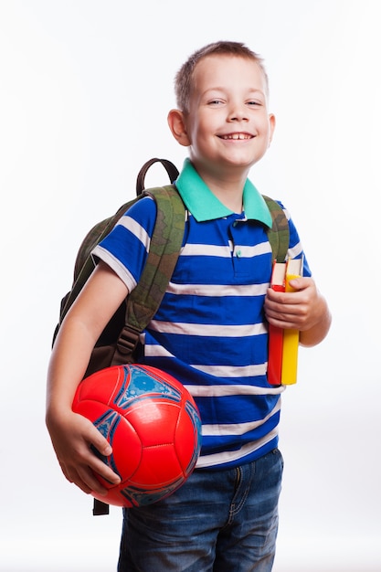 Colegial feliz con la mochila, la bola y los libros aislados en el fondo blanco