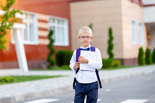 Un colegial con gafas rubias con una mochila y un libro blanco está de pie en la escuela .. Día del conocimiento