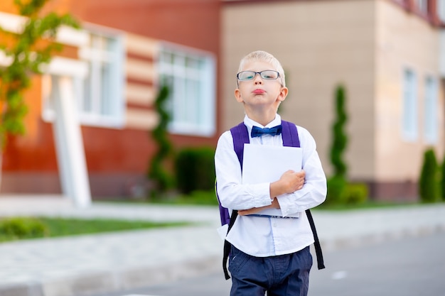 Un colegial con gafas rubias con una mochila y un libro blanco está de pie en la escuela.