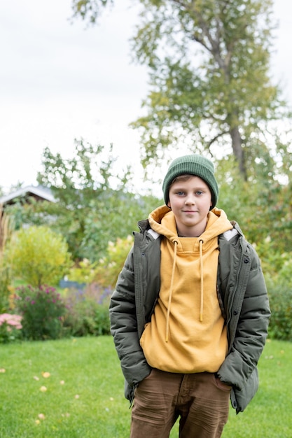 Colegial guapo y adorable en gorro, sudadera con capucha, pantalones y chaqueta de abrigo de pie sobre el césped verde frente a la cámara en el jardín