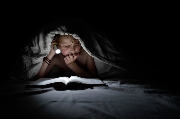 Colegial leyendo debajo de la manta por la noche.