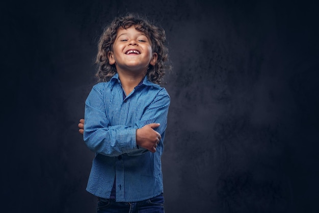 Un colegial lindo que se ríe con el pelo rizado castaño vestido con una camisa azul, posando en un estudio. Aislado en el fondo oscuro con textura.