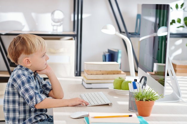 El colegial pensativo sentado a la mesa en casa y viendo la lección en línea en la pantalla de la computadora mientras educa en el hogar