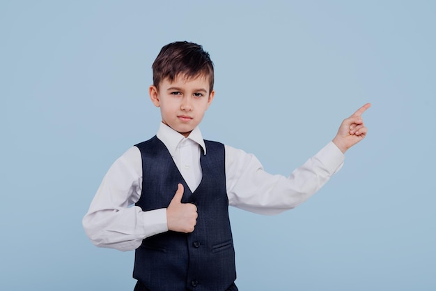 Colegial pequeño serio en camisa y chaleco apuntando con el dedo y mostrando el pulgar hacia arriba en el estudio en blu