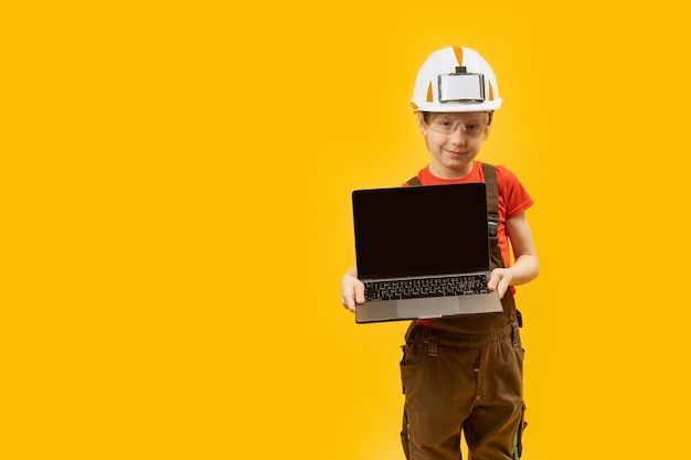 Colegial en ropa de trabajo y casco protector sostiene portátil con pantalla vacía Niño como arquitecto Elección de futura profesión Copiar espacio