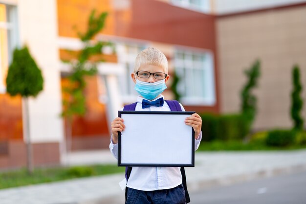 Un colegial rubio con gafas y una máscara protectora se encuentra en la escuela y sostiene un cartel con una sábana blanca. Día de los conocimientos.