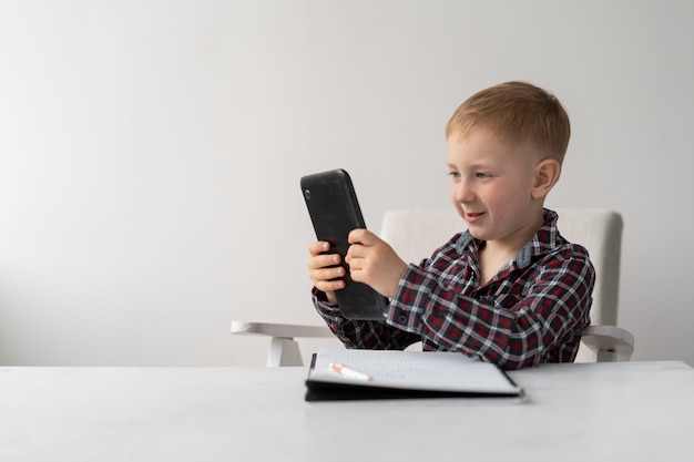 El colegial rubio se sienta en una silla frente a la mesa. Aprendizaje a distancia y cuarentena. El niño tiene una tableta en sus manos y un cuaderno para escribir sobre la mesa.