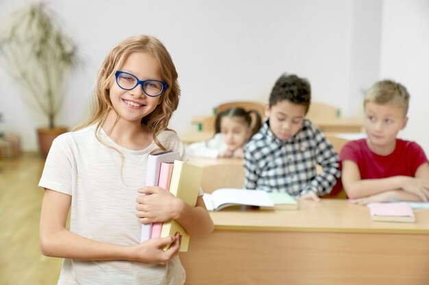 Colegial segurando livros, em pé na sala de aula, sorrindo.