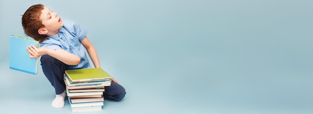 Colegial sentado con un montón de libros escolares y tirando un libro de texto aislado en un fondo azul