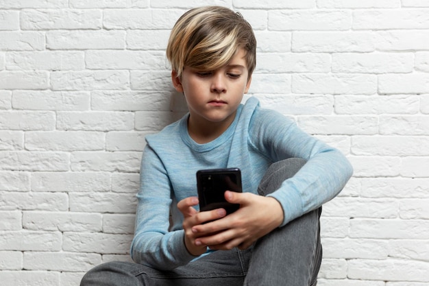 Un colegial se sienta con un teléfono en las manos contra el fondo de una pared de ladrillos blancos Un chico guapo con un suéter azul y pantalones grises Juegos de chat y redes sociales