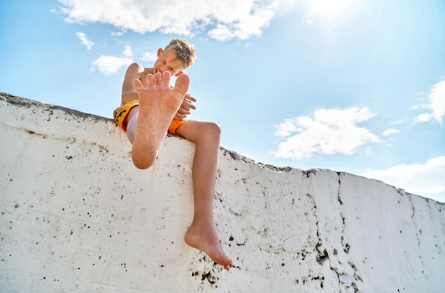 Un colegial sonriendo alegremente en un muelle alto y levantando el pie y disfrutando de las vacaciones de verano junto al mar