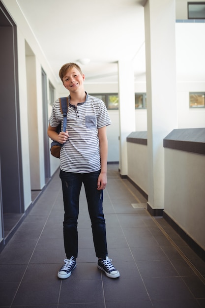 Foto colegial sonriente de pie con mochila en el pasillo de la escuela