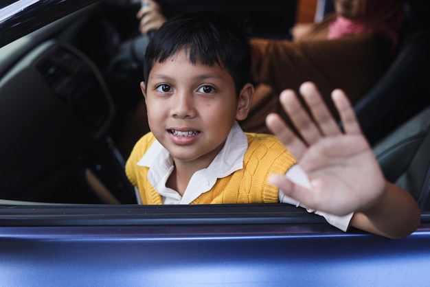 Un colegial sonriente con uniforme agita la mano desde la ventana del auto listo para ir a la escuela