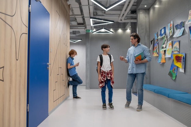 Foto colegial con el teléfono inteligente apoyado contra la pared mientras su alegre compañero de clase se comunica con un maestro serio