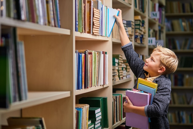 colegial tomando libros de los estantes de la biblioteca, con una pila de libros en las manos. desarrollo del cerebro infantil, aprender a leer, concepto de habilidades cognitivas