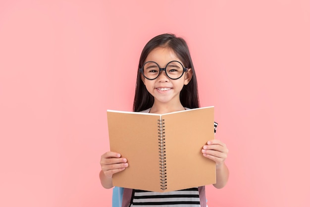 Colegiala abrazando libro vistiendo mochila sonriendo aislado sobre fondo de color rosa