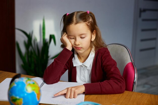 Una colegiala aburrida se sienta en el escritorio haciendo la tarea leyendo educación en el hogar