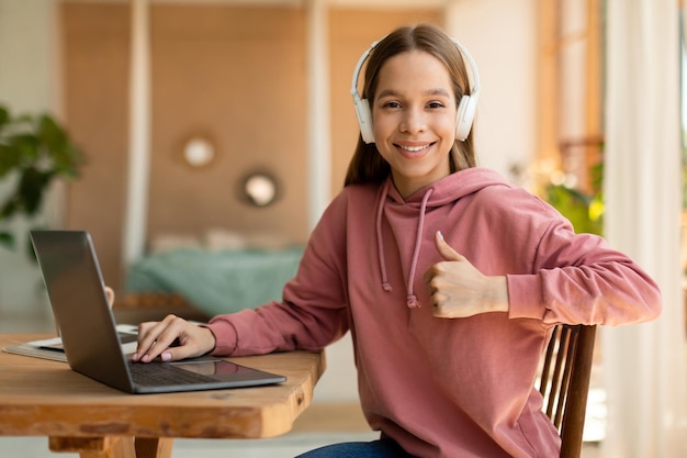 Foto colegiala adolescente feliz en auriculares gesticulando con el pulgar hacia arriba sentado en el escritorio cerca de la computadora portátil que tiene una lección en línea desde casa