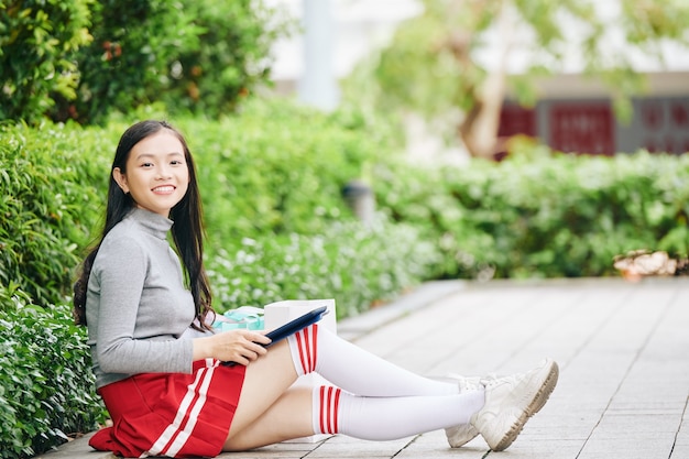 Colegiala adolescente feliz sentado en el suelo y usando la aplicación en la computadora de la tableta