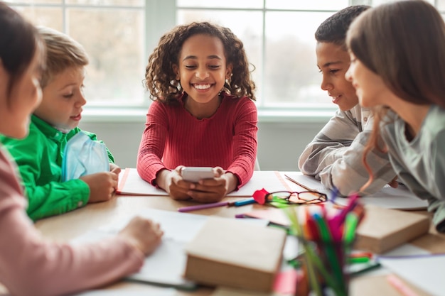 Colegiala africana feliz usando teléfono sentado con compañeros de clase en el aula